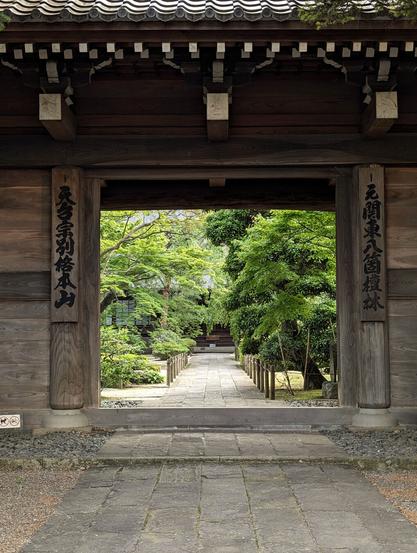 A temple garden and green trees can be seen through a big, open japanese-style gate.