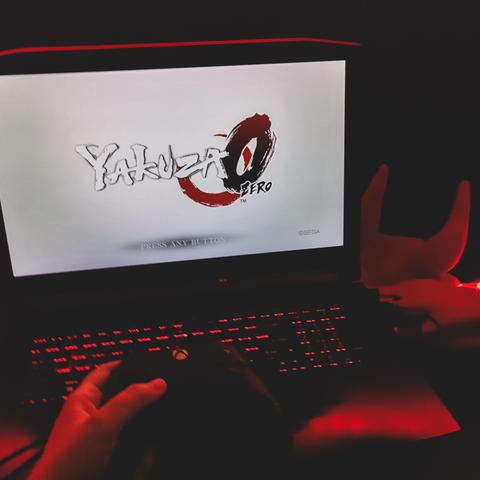yakuza 0 zero video game main menu start scree, white with red zero and crescend in the background, font imitating brush strokes. video game is playing on a hellios 300 predator (asus) laptop, red lit keyboard, and red lit room, with balaclava with devil horns on it in the corner, and a hand holding xbox controler in the foreground, hovering above the keyboard. review and picture by wannabekingpin aka nosfeartu reads