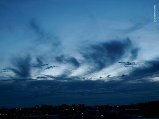 A dark blue cloud bank on the horizon completely obscures a coming sunrise. Higher up the sky is clearer, with only gauzy, wispy clouds that are also a deep blue. Behind, the sky is a much lighter blue, but there is no sign of sunrise colours. Underneath the clouds the distant land is completely dark, but given shape by many lit windows and streetlights.