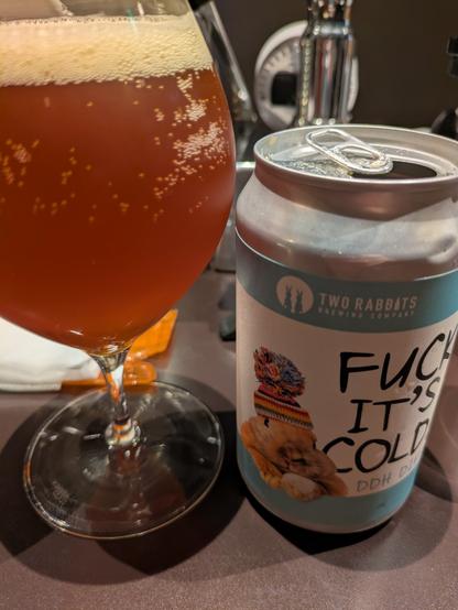 A glass filled to the brim with beer. The empty can stand next to it, the label reads "Fuck It's Cold IPA"