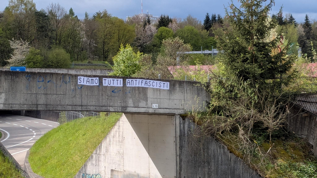 A pedestrian overpass with a feminist collage pasted on it that reads: SIAMO TUTTI ANTIFASCISTI. On the left a street and in the background many blooming trees.