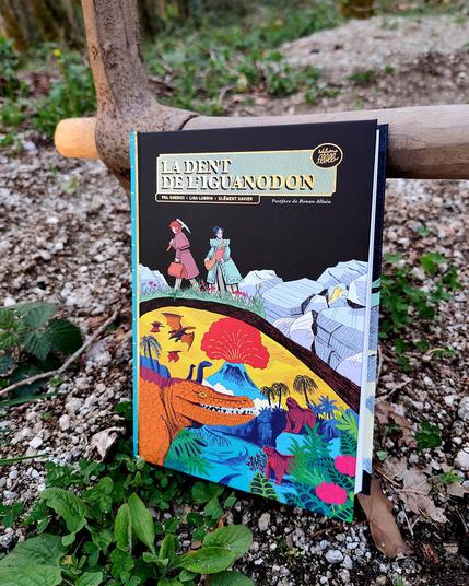 Photo de la BD "La dent de l'iguanodon" de Lisa Lugrin, Clément Xavier et Pol Cherici, sur un sol caillouteux dans lequel est planté une pioche (subtile allusion à la couverture et à la vie de ces paléontologues du XIXeme)