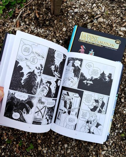 Photo de la BD "La dent de l'iguanodon" de Lisa Lugrin, Clément Xavier et Pol Cherici, sur un sol caillouteux dans lequel est planté une pioche (subtile allusion à la couverture et à la vie de ces paléontologues du XIXeme) Livre ouvert sur une double page.