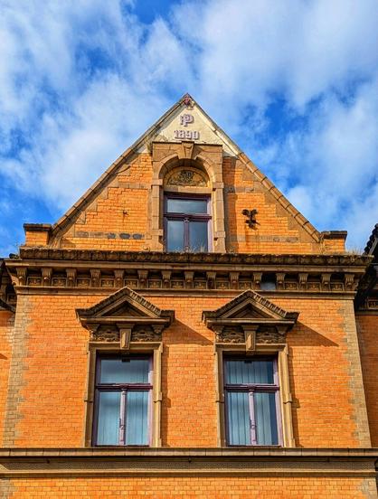 A low-angle shot showcases the detailed brickwork of a historic building against a vibrant blue sky with scattered white clouds. The building is constructed from orange-red bricks with lighter, sandy-coloured brick accents forming decorative patterns around the windows and along the roofline. Two prominent rectangular windows with multiple panes and light-coloured frames are visible on the lower level. Above, a smaller, arched window with similar framing is centred within the triangular gable. A decorative stone or brick element featuring the year "1890" is positioned above the arched window. The roof is tiled in a darker reddish-brown. The architectural style suggests a late 19th-century construction.