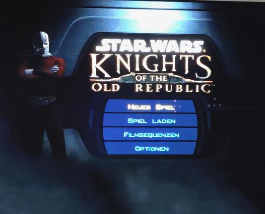 Der (schlecht abfotografierte) Startbildschirm von "Knights of the Old Republic".
Im Zentrum steht das Hauptmenü, "Neues Spiel" ist ausgewählt.
Darüber ist groß der Titel-Schriftzug und links steht ein Mann mit tätowierter Glatze und einer Art Maske über der Mundpartie.
Zu seinen Füßen wabert Nebel.