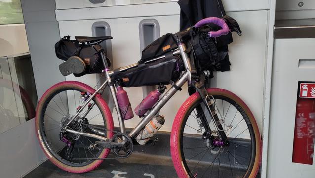 Vélo gravel en titane avec accessoires violets, pneus roses et bagagerie Restrap. Il est posé devant les crochets porte vélo inadaptés d'un TER