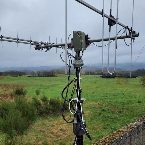 Das Bild zeigt eine große Antenne, die auf einem Mast in einer ländlichen, grünen Landschaft montiert ist. Die Antenne besteht aus mehreren Elementen, darunter Yagi-Elemente und eine U-förmige Schleife. Am Mast befinden sich verschiedene Kabel, die lose hängen, sowie ein wettergeschütztes Gehäuse, vermutlich für die Steuerung oder Verstärkung der Antennensignale. Im Hintergrund sind grüne Felder, Büsche und weiter entfernte bewaldete Hügel zu sehen. Der Himmel ist bewölkt und grau, was auf ein regnerisches oder feuchtes Wetter hindeutet. Man erkennt auch einige Windräder in der Ferne.