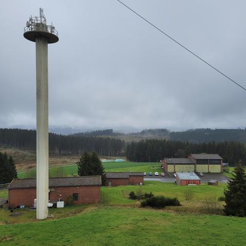 Das Bild zeigt ein weitläufiges, eingezäuntes Gelände in ländlicher Umgebung mit mehreren Backsteingebäuden und einem markanten, hohen Betonturm auf der linken Seite. Auf dem Turm befinden sich diverse Antennen und technische Einrichtungen, was auf eine Funk- oder Telekommunikationsnutzung hindeutet.

Im Hintergrund sind dichte Nadelwälder und leicht nebelverhangene Hügel zu sehen, was auf eine höher gelegene, feuchte Region schließen lässt. Auf dem Gelände parken einige Fahrzeuge, was auf eine aktive Nutzung oder Wartung des Standorts hindeutet. Insgesamt wirkt die Anlage wie eine technische Betriebsstelle, möglicherweise für Kommunikation, Rundfunk oder militärische Zwecke.