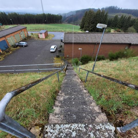 Das Bild zeigt eine steile Betontreppe mit Metallgeländern, die einen grasbewachsenen Hang hinabführt. Am unteren Ende der Treppe befinden sich mehrere Gebäude mit roten Ziegelwänden und Flachdächern, vermutlich Funktions- oder Betriebsgebäude. Auf dem gepflasterten Hof davor stehen einige Autos, was auf eine aktive Nutzung hindeutet. Die Umgebung ist von dichten Wäldern und grünen Wiesen geprägt, typisch für eine ländliche, hügelige Region. Der Himmel ist bedeckt und es scheint feucht zu sein, was das moosige und verwitterte Aussehen der Treppe erklärt. Im Hintergrund ist ein Windrad zwischen den Bäumen zu erkennen.