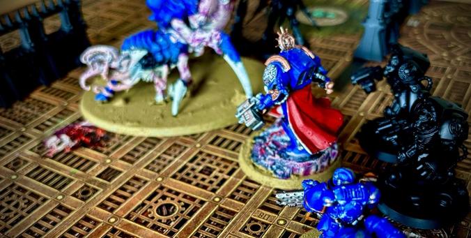 Das Bild zeigt eine detaillierte Szene aus Warhammer 50k - einem Tabletop-Spiel, bei dem mehrere Miniaturfiguren auf einem kunstvoll gestalteten Spielfeld positioniert sind. Das Spielfeld hat ein industrielles Design mit einem Gittermuster und mechanischen Elementen, was auf eine futuristische oder technologisch geprägte Umgebung hinweist. Im Hintergrund eine sogenannte Psychophage der Tyraniden, vorne Ultramarines.