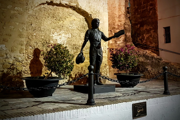 Vor einer alten Stadtmauer steht die Statue eines Mannes. In der einen Hand hält er gesenkt seinen Hut. In der anderen erhoben eine Art Urkunde.