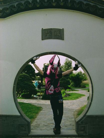 A fursuiter standing in the middle of a circular passage in the a wall. The fursuiter wears a pink skull mask with pink and white hair and black and pink ears. They hold there arms up to the circle. The wall is plastered white with a darker first row of stone. Above the passage there is the name "Huarui" (a welding equipment manufacturer) in Chinese characters.