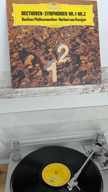 Das Bild zeigt ein klassisches Schallplattencover und einen Plattenspieler mit aufgelegter Vinyl-Schallplatte. Oben ist das Cover der Aufnahme zu sehen:

Es handelt sich um eine Schallplatte der Deutschen Grammophon mit den „Symphonien Nr. 1 und Nr. 2“ von Ludwig van Beethoven.

Gespielt von den Berliner Philharmonikern, dirigiert von Herbert von Karajan.

Auf dem Cover sind große, stilisierte Ziffern „1“ und „2“ dargestellt, die in einer felsigen Landschaft stehen.

Im unteren Teil des Bildes sieht man den laufenden Plattenspieler, auf dem die schwarze Vinylplatte mit gelbem Etikett liegt. Der Tonarm ist aufgelegt, die Musik wird also gerade abgespielt. Das Gesamtbild vermittelt ein Gefühl von klassischer Musikleidenschaft und analogem Musikgenuss.