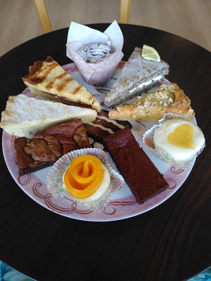 Auf einer rosa-weißen Kuchenplatte thronen neun verschiedene Kuchenstücke (im Uhrzeigersinn): 
Bananenkuchen mit Pekannüssen (in einem Papierförmchen mit hohem Rand gebacken und mit Puderzucker bestäubt), Zitronen-Mohn-Kuchen (mit einem Puderzuckerguss, einer Viertelscheibe Zitrone als Deko sowie einer Frischkäsecrème als Füllung), eine Birnen-Mandel-Tarte (mit Mandelblättchen bestreut), ein Cheesecake mit Lemon Curd (mit einem eingebackenen Herzen aus dieser fantastischen Zitronencrème), ein längliches, rechteckiges Stück klassischer Brownie (wunderbar schokoladig und so weich wie eine gebackene Praline), ein Mango Cheesecake (die Mango sitzt, in Scheiben geschnitten, als Rose auf dem Cheesecake, der mit Vanillemark parfümiert ist), ein längliches, rechteckiges Stück Brownie mit eingebackenem Schokokeksteig, ein dreieckiges Stück Shortbread mit Tonkabohne (mit etwas Zucker bestreut) sowie ein dreieckiges Stück Shortbread mit Karamell (dekoriert mit einigen Streifen Karamell).