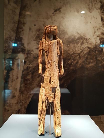 Photograph of the Löwenmensch (Lion-man) figurine, a sculpture carved from mammoth ivory. The figure stands upright with a humanoid body and a lion-like head, showing a blend of human and animal features. The surface is weathered and fragmented, with visible cracks and missing sections. It is displayed in a museum setting, with a blurred, textured background resembling stone or cave walls.