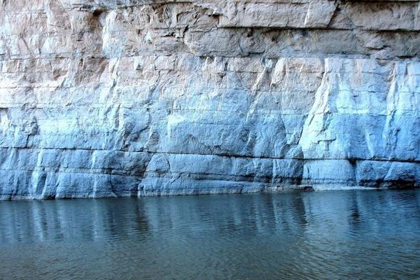 A rocky cliff face in Big Bend National Park rises above the calm water of the Rio Grande. Layers of stone in shades of gray and brown reveal textures and cracks. The still water below reflects the serene majesty of the cliff, appearing completely undisturbed.