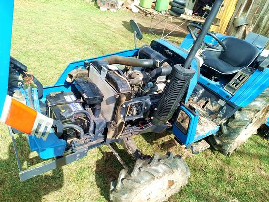 Front of the same tractor, with the engine panels removed to expose a small 3 cylinder diesel engine, battery, radiator and alternator with new belt installed.