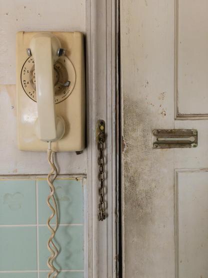An old rotary telephone with a long, coiled cord, mounted on a dirty wall next to a dirty door with an unlocked chain latch.