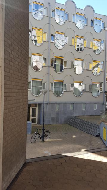 A black bicycle parked against a single lamppost on a small paved square in front a five story building. The ground floor has a white framed entrance door and small vertical oriented rectangular windows in a dark  grey stone facade. 

The other floors have light grey painted facade withrows of shield shaped = a square with a half circle on the bottom = windows with a yellow top accent, flanked by small, rectangular window on either side. The space between the windows have mirrored accents = a half circle on top of a square =  give the facade a wave like appearance.

Most of the windows show yellow and white reflections of pieces of out of frame buildings.