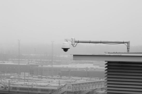 A black and white picture of a 360 surveillance camera mounted at the end of a white metallic arm, on the edge of a roof. In the distance a train exchange in the fog.