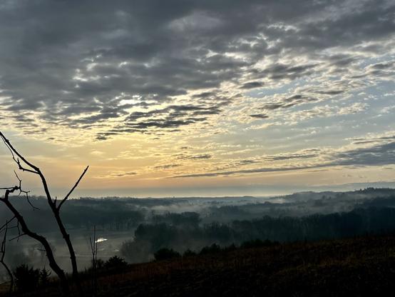 View from a small hilltop overlooking a valley partially filled with fog. Small groves of trees poke through the fog. A creek meanders through the valley. Early morning sunlight glints off its surface. The sun is blocked by the last remnants of clouds from a storm.