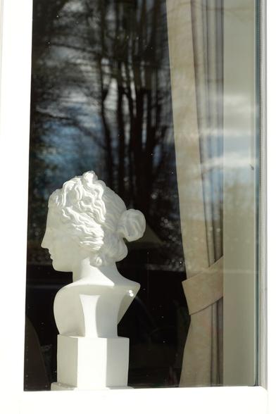 Blick durch ein Fenster auf eine Büste einer Frau, die schräg in den Raum schaut. Rechts am Fenster ist ein heller Vorhang, der mit einem Band zusammengefasst ist. In der Scheibe spiegelt sich der blaue, bewölkte Himmel und unbelaubte Bäume.