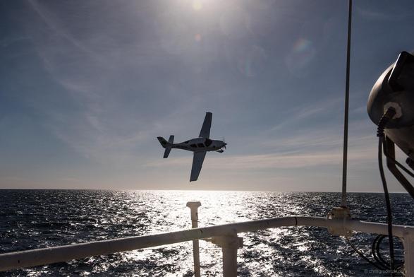 Ein kleines Propellerflugzeug im Gegenlicht über dem Meer, im Vordergrund eine Schiffs-Railing.