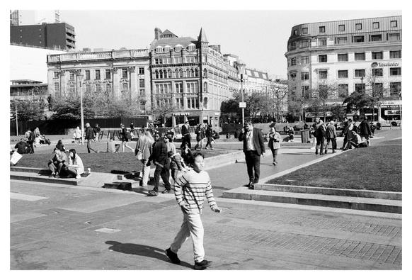 The image showcases a bustling urban park with people engaged in various activities. In the foreground, a kid in a striped shirt and light-colored pants walks toward the camera, adding a dynamic element to the scene. Behind him, the park is filled with individuals and groups sitting on the grass, walking, and interacting.
The background features a mix of modern and historic architecture, including a prominent building with ornate details and a spire, which adds depth and context to the location. The sky is clear, suggesting a bright and sunny day, which illuminates the scene and casts shadows that enhance the texture and dimension of the photograph.