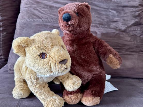 A stuffed lion and a stuffed brown bear are sitting together on a brown couch. The lion is light tan with orange eyes, while the bear is dark brown with a black nose.