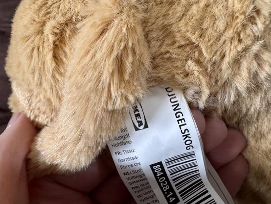 A close-up of a fluffy, light brown stuffed lion cub, being held in a hand. A tag with multilingual information is visible, indicating the product name "DJUNGELSKOG" from IKEA.