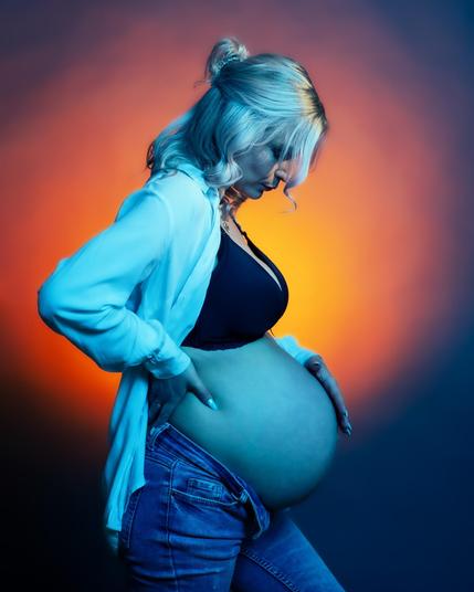 The image shows a pregnant woman standing in profile with a hand on her back and the other on her belly. The background is a gradient of orange and blue lighting.