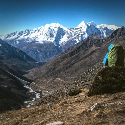 A sitting person on a rock with a backpack looking at a snowy mountain panorama.