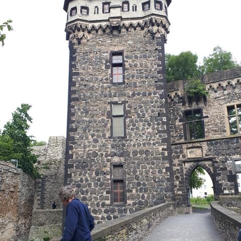 Das Bild zeigt eine mittelalterliche Steinburg mit einem hohen, zylindrischen Turm, der aus dunklen und hellen Steinen gebaut ist. Der Turm hat mehrere Fenster mit schmiedeeisernen Gittern und ist mit gotischen Verzierungen an der Spitze versehen. Neben dem Turm ist ein Torbogen zu sehen, der ebenfalls aus Stein gefertigt ist und mit einem steinernen Wappen verziert ist. Der Weg vor dem Torbogen führt zu einem gepflasterten Bereich, auf dem ein Person mit grauem Haar und einem blauen Pullover spazieren geht. Im Hintergrund sind Bäume und ein Teil eines modernen Gebäudes zu erkennen. Die Burg ist von einer Mauer umgeben, die teilweise von Pflanzen überwuchert ist.

Bereitgestellt von @altbot, privat und lokal generiert mit Ovis2-8B
