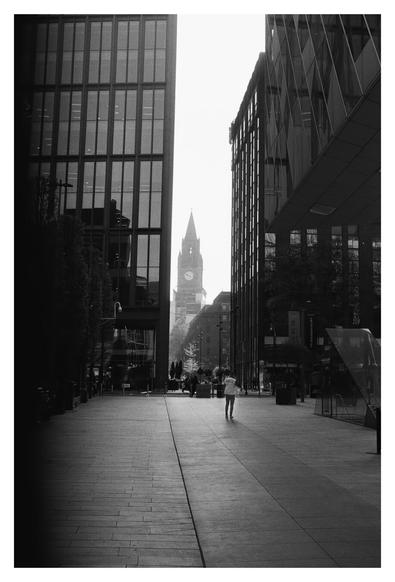 This black-and-white photograph captures a quiet urban scene. The viewer stands on a wide, empty sidewalk flanked by tall, modern buildings with large glass windows. The buildings frame a distant clock tower, which stands prominently in the center background, its spire reaching into a clear sky. A solitary figure walks away from the camera down the sidewalk, adding a sense of scale and life to the scene. The overall atmosphere is serene and slightly moody, with the stark contrast between the dark buildings and the bright, open space creating a dramatic effect.