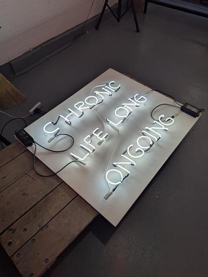 An Abbey Divens piece dealing with chronic illness and medication. Her handwriting is a little sloppy with some crazy bends and blown out powders- which in this case makes the piece stronger. White neon letters reading "CHRONIC LIFE LONG ONGOING" mounted on a white rectangle with two black, visible transformers on the face.
