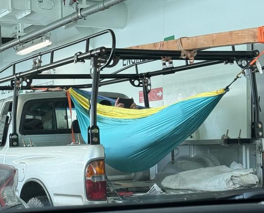 A person in a hammock in the back of a pickup truck aboard a ferry, the hammock is hung from a large piece of wood on the roof rack