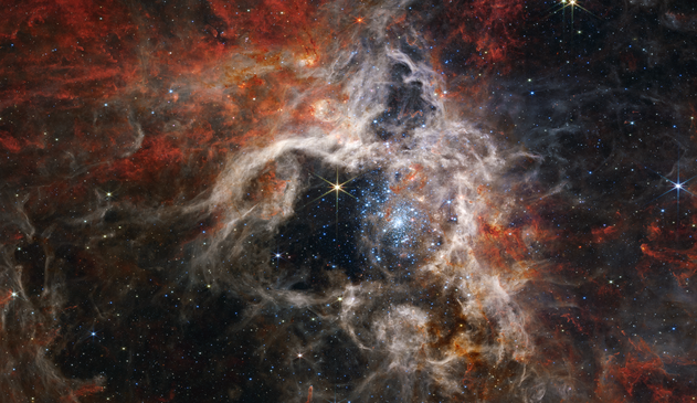 A star-forming nebula. The nebula is composed of tan-colored clouds with rust-colored highlights, surrounding a black central area containing a bright cluster of sparkling pale blue stars that scatter outward from a densely packed center. The clouds toward the center are brighter than those toward the edge. The bottom left and the top right of the nebula appear more clear of any clouds of dust and gas. One bright yellow star stands out in the central open area, showing off Webb’s eight-pronged diffraction pattern.