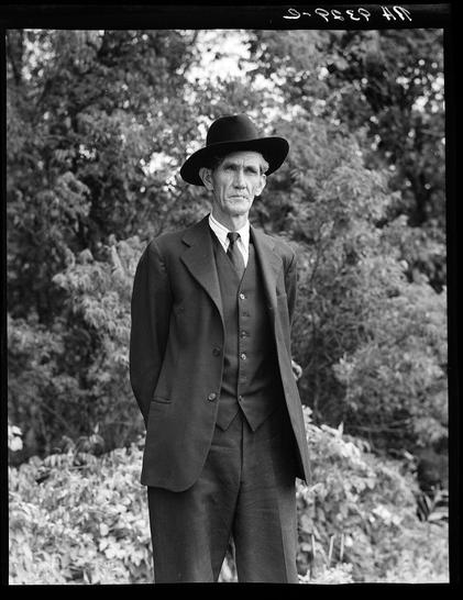 The image is a black and white photograph of an elderly man standing in what appears to be a garden or park. The man is wearing a dark suit, vest, tie, and a wide-brimmed hat, which gives him the appearance of someone from early 20th-century America. He has a stern expression on his face as he looks directly at the camera. Behind him are trees and bushes that suggest a natural outdoor setting.

The photograph is labeled with handwritten text in white ink along the top edge: "S.-PETER HEN." The handwriting appears to be part of an archival collection, possibly from Dorothea Lange's famous series capturing life during the Great Depression or other historical moments. This particular image seems to evoke a sense of rural American heritage and is reminiscent of iconic portraits taken in that era.

Additional information about this photo can likely be found within archives associated with historic photographs depicting societal conditions of the time, such as Dorothea Lange's work for the Farm Security Administration (FSA) during the Great Depression.