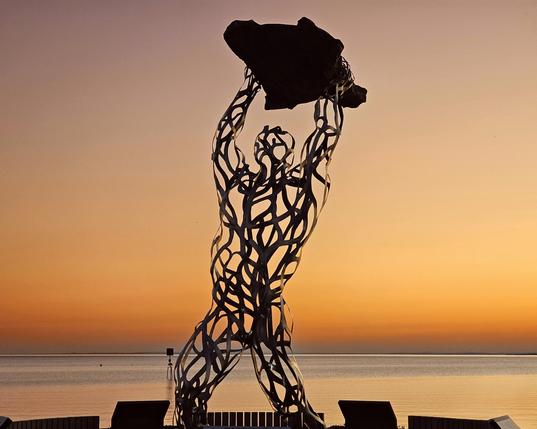 A tall, abstract metal sculpture of a human-like figure lifting a dark, irregular shape above its head, silhouetted against a vibrant orange and purple sunset over a calm body of water.
