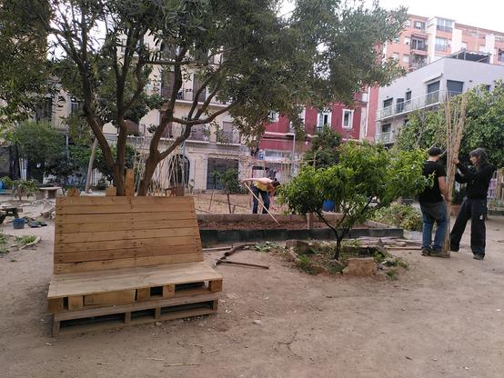 Foto de l'horta. Es veu un banc i un arbre a l'esquerra, un arbre petito i dues persones agafant canyes a la dreta i una persona al centre preparant un bancal.