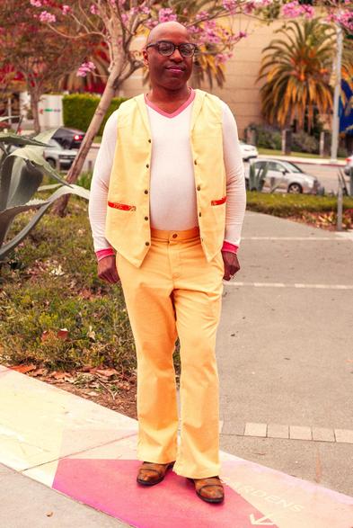 A man with a shaved head and glasses poses in an urban park with blooming Hong Kong orchid trees with their pink blossoms in the background. He wears matching butter yellow vest and pants with orange satin brocade details on the pockets over a fitted, long sleeve t-shirt made of blush pink with raspberry cuffs and collar.