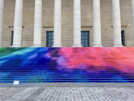 Une partie de la façade nord du Palais Bourbon, quai d'Orsay. 
Au pied des douze colonnes, les contremarches de l'escalier sont couvertes de lattes de bois multicolores : vert, bleu, mauve, rose, orange.