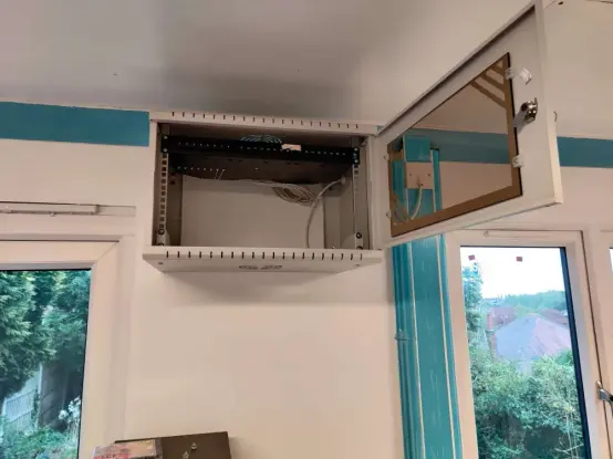 A small comms cabinet that looks a bit like a microwave when closed is bolted high up on a white wall. Inside is the 24 socket patch panel from previous picture.