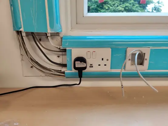 Office dado trunking coming down a wall and turning 90 degrees to the right. The corner panel is removed, showing the separate areas for data cables and power cables.  There is a plug socket on the trunking as well as an empty network socket with a couple of cables poking through.