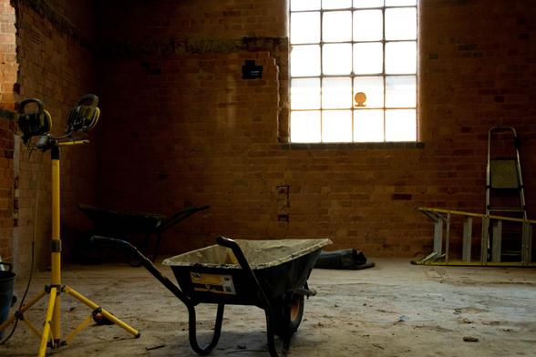 A weelbarrow, a tripod with lights, and two ladders in a gutted space.