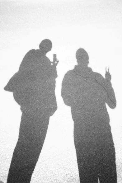 A black and white photo of just the shadows of two people. The person on the left can be seen holding the camera while the person on the right is giving a peace sign.