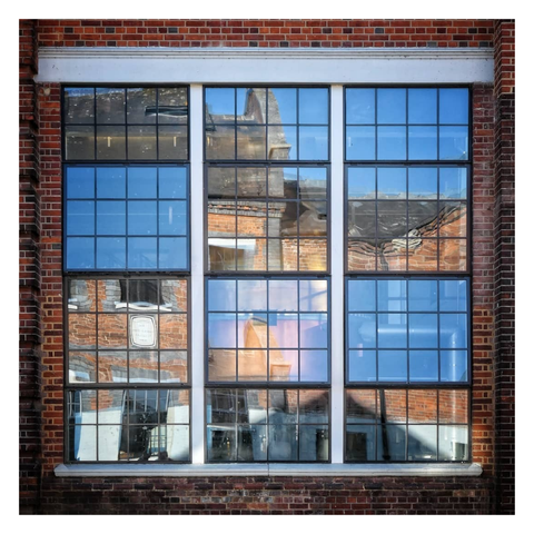 This is a square format colour image of a square window comprising multiple glass panes, and surrounded by brickwork, with a white lintel at the top. There are three panels of 2 by 4 panes to the top and bottom, and two rows of 3 by 4 panes to the middle. Some of the panels are propped open, reflecting the blue sky, while others are closed and reflect either the brickwork of the building opposite or show glimpses of the copper colour of a large gin still which sits behind the window. The effect creates an abstract image of shades of bronze and blue across a grid. Taken at the Bombay Sapphire distillery with a Sony RX100.