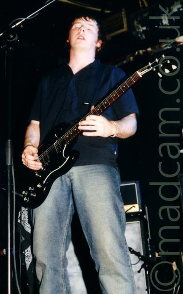 Low view of a man with dark hair, wearing a black shirt and a faded blue pair of jeans, playing a black electric guitar.
He is leaning back slightly, with his eyes closed and his mouth slightly open.
Black and grey amplifiers line the black walls in the background, with the ceiling also being black.