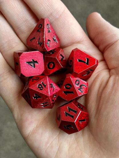 Picture of a set of red polyhedral dice with black numbers and a shiny metallic finish, sitting in a hand.