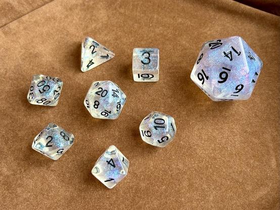 Picture of a set of transparent polyhedral dice with black numbers & blue-purple glitter inside, including an extra-large d20, sitting on a brown felt background.